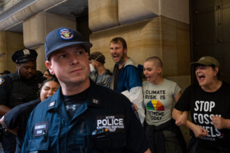 Climate protesters block the doors to the Federal Reserve Bank of New York on Monday as an NYPD police officer with the strategic Response Group, which specializes in large demonstrations, crowd control, and major events, center, watches over the demonstrators and another officer arrests a protester, left. Credit: Keerti Gopal